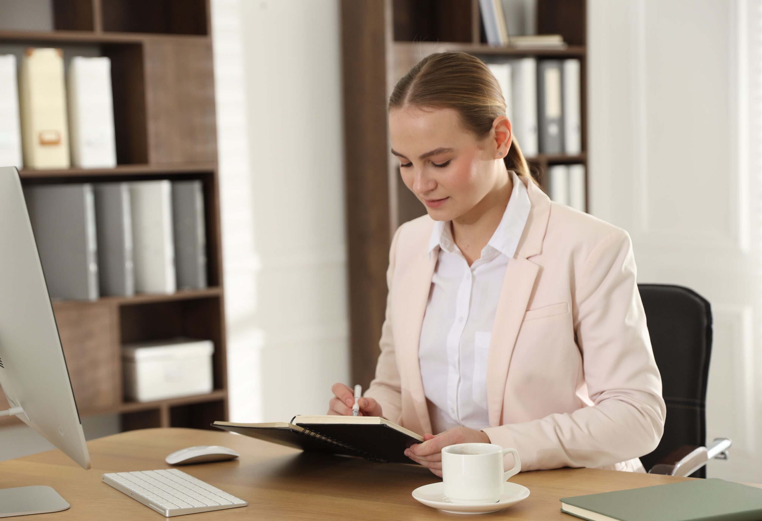 Professional woman taking notes at her desk beside a computer and coffee.