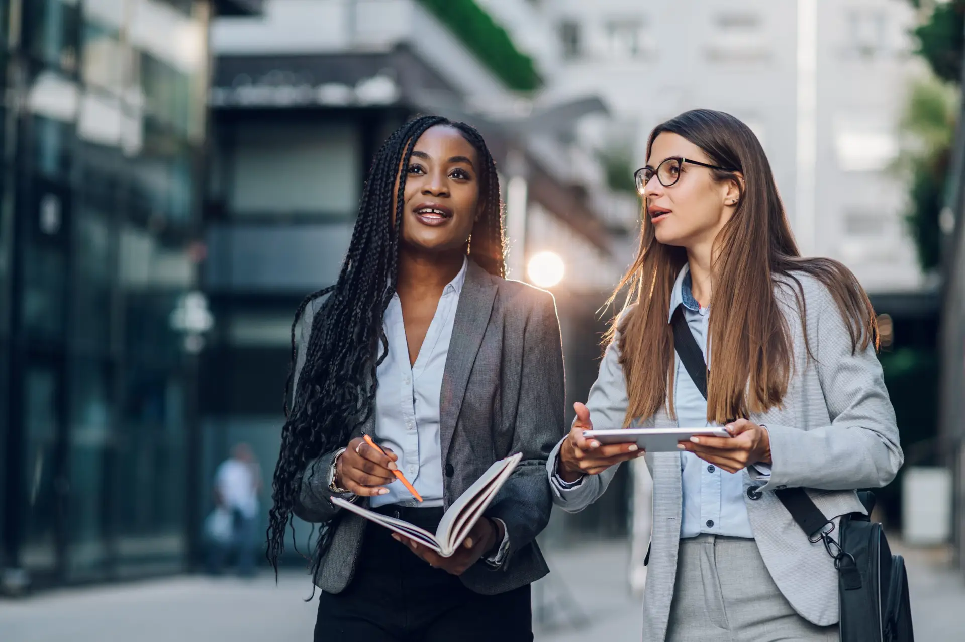 Two women in business attire talk outdoors, one holding an open notebook and pencil and the other a tablet.