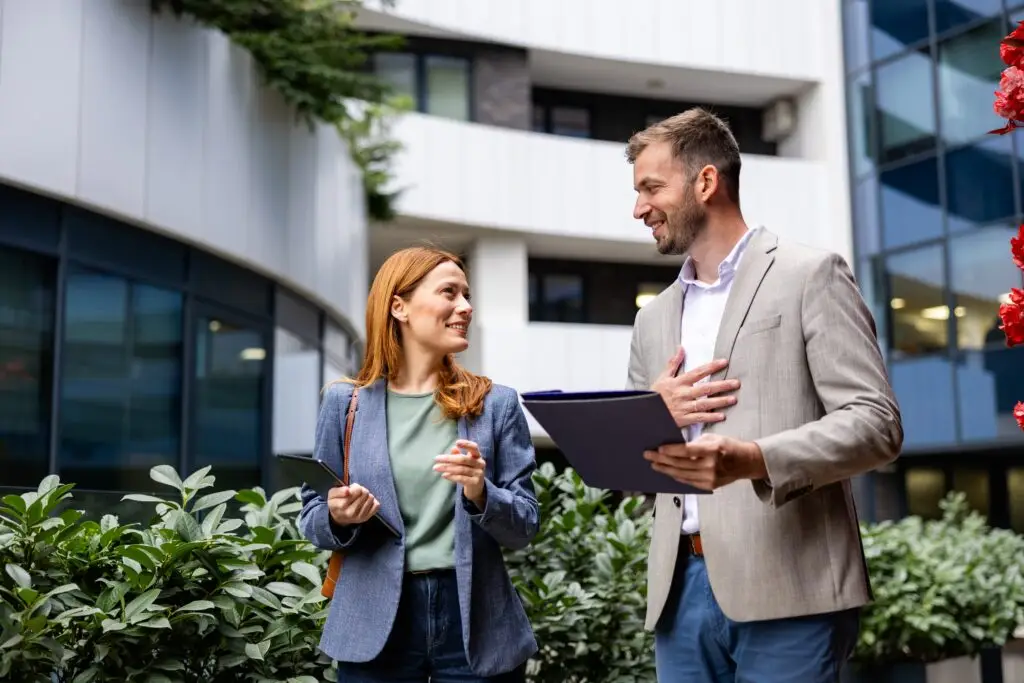 A woman holding a tablet and a man holding an open folder smile and talk outside a modern building.