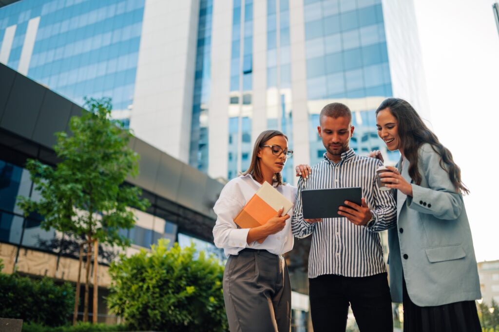 Three people gather around a tablet outside an office building; one woman holds folders and another holds a takeaway cup.