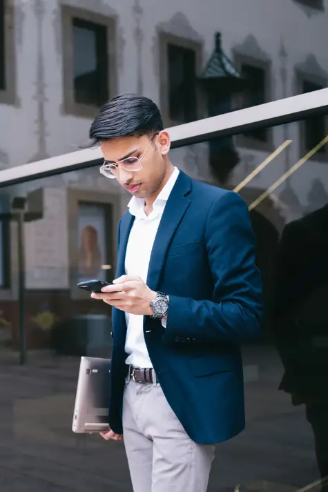A man in a blue blazer checks his smartphone while carrying a laptop beside a reflective glass building.